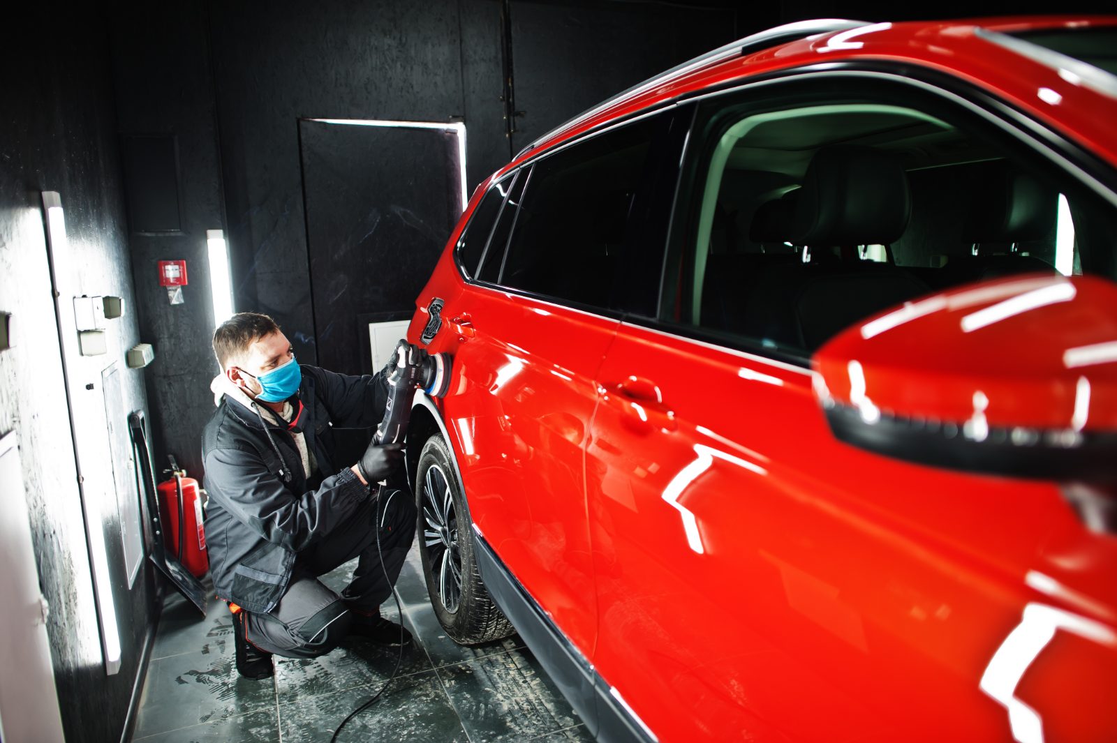 Pozycjonowanie auto detailingu - detailer w maseczce poleruje polerką orbitalną karoserię czerwonego SUV-a w studiu