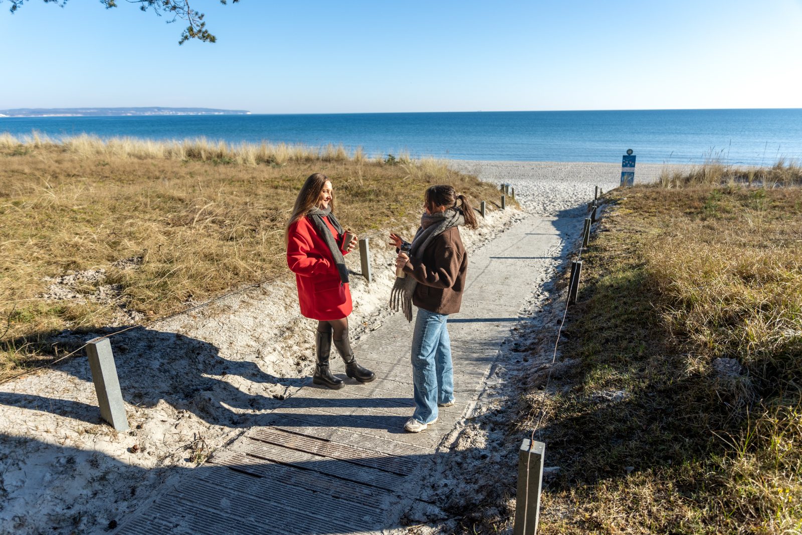Pozycjonowanie biur turystyki krajowej - dwie kobiety w ciepłych kurtkach na ścieżce przez wydmy prowadzącej na plażę nad morzem