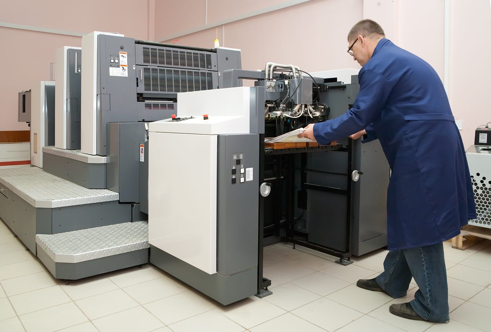 Pozycjonowanie drukarni - operator w niebieskim fartuchu obsługuje maszynę offsetową w hali drukarni