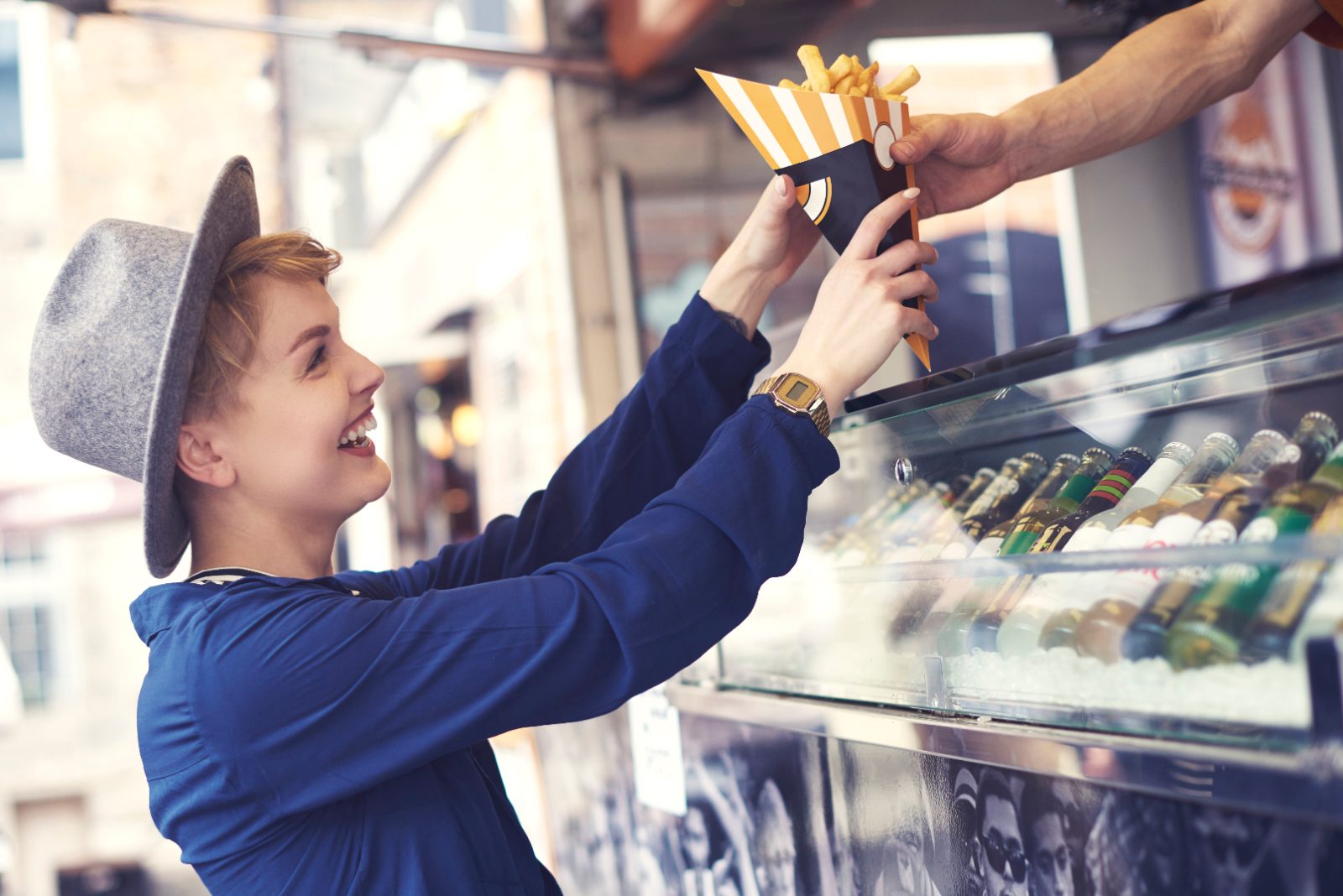 Pozycjonowanie food trucków - uśmiechnięta klientka w kapeluszu odbiera frytki z okienka food trucka, obok witryna z napojami