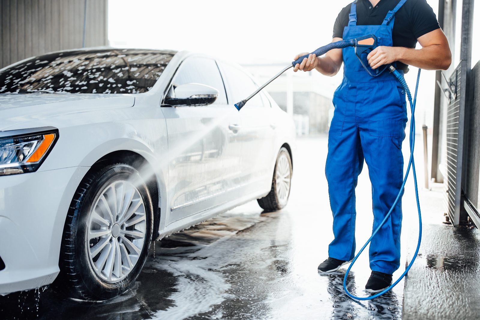 Pozycjonowanie myjni samochodowej - pracownik w niebieskim kombinezonie myje białe auto myjką ciśnieniową