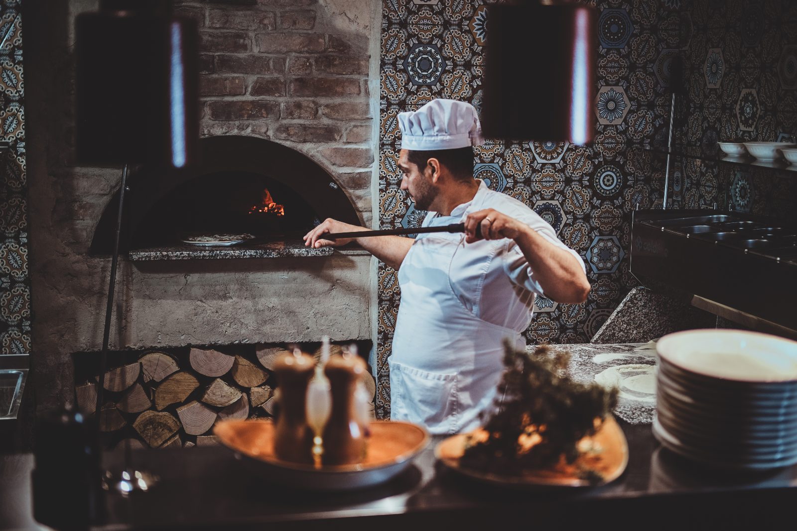 Pozycjonowanie pizzerii - pizzaiolo w białej czapce wkłada pizzę łopatą do pieca opalanego drewnem, obok ułożone polana