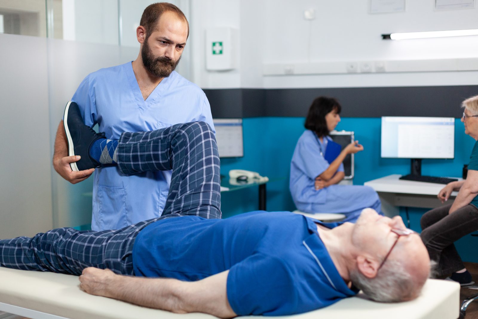 Pozycjonowanie rehabilitacji - fizjoterapeuta ćwiczy nogę starszego pacjenta leżącego na kozetce w sali rehabilitacyjnej